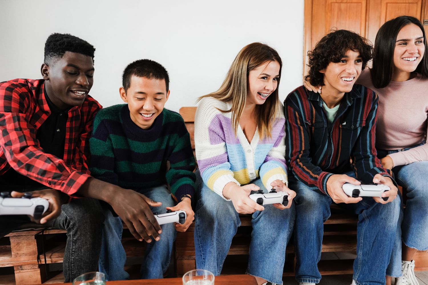 A diverse group of five young friends sitting side-by-side on a wooden pallet bench, laughing and smiling as they play a multiplayer video game together with white controllers.